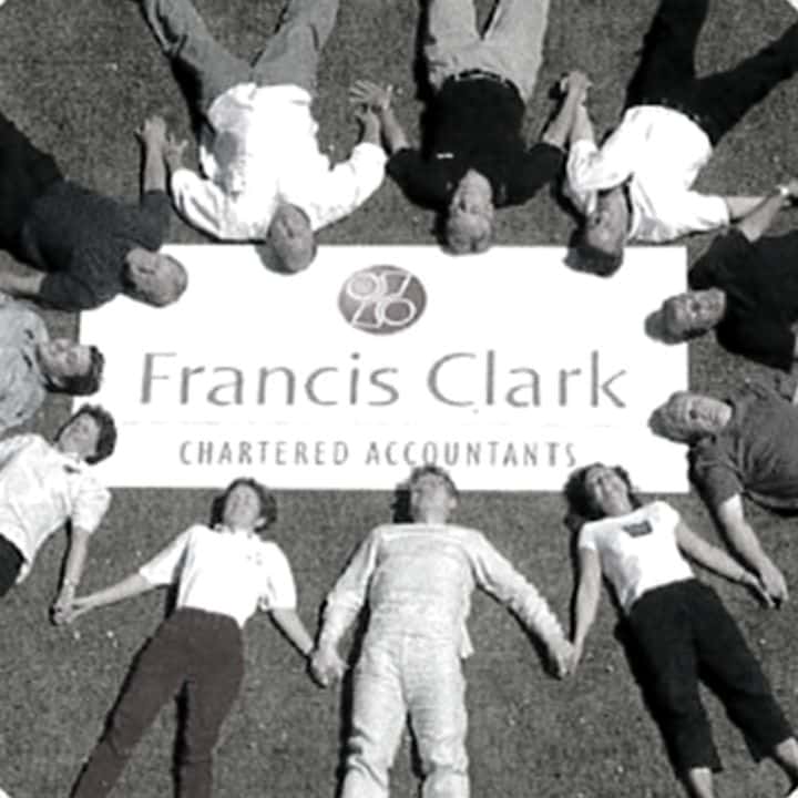 Group of colleagues lying on the ground in a star shape smiling surrounding a Francis Clark sign