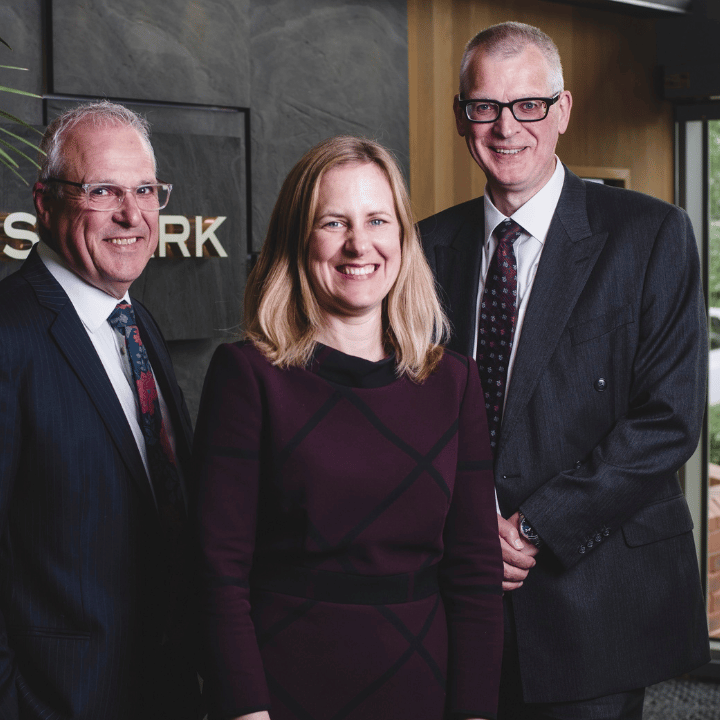 Three smartly dressed professionals smiling at the camera in front of a slate wall