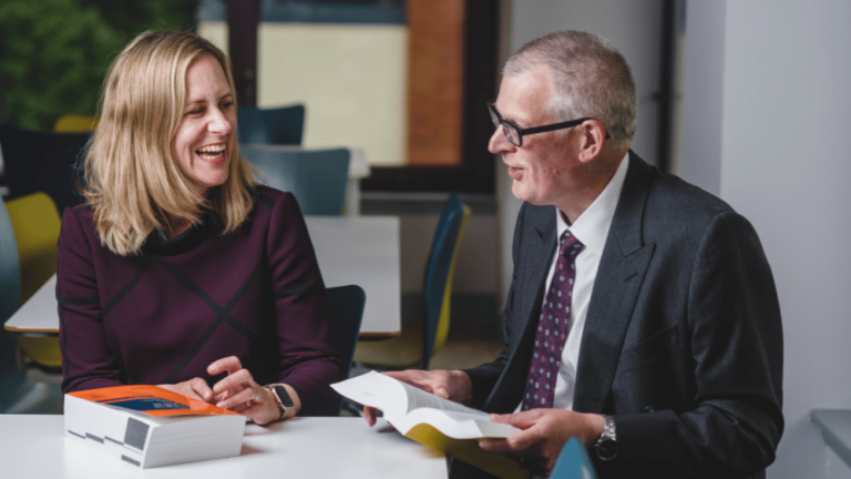 A male and female are seated at a desk with two reference books engaged in a conversation.