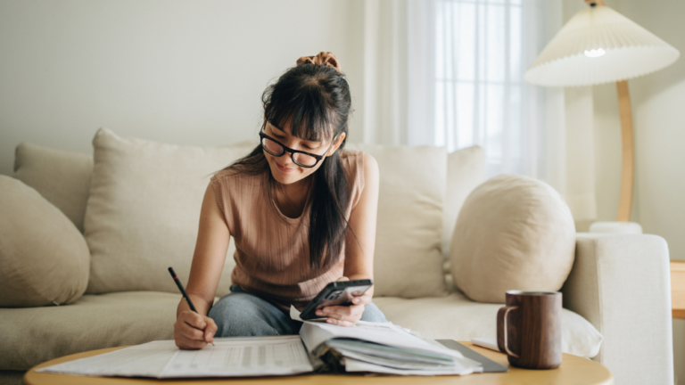 Young female sitting on a settee in her living room filling out forms on a coffee table with a phone in her hand.